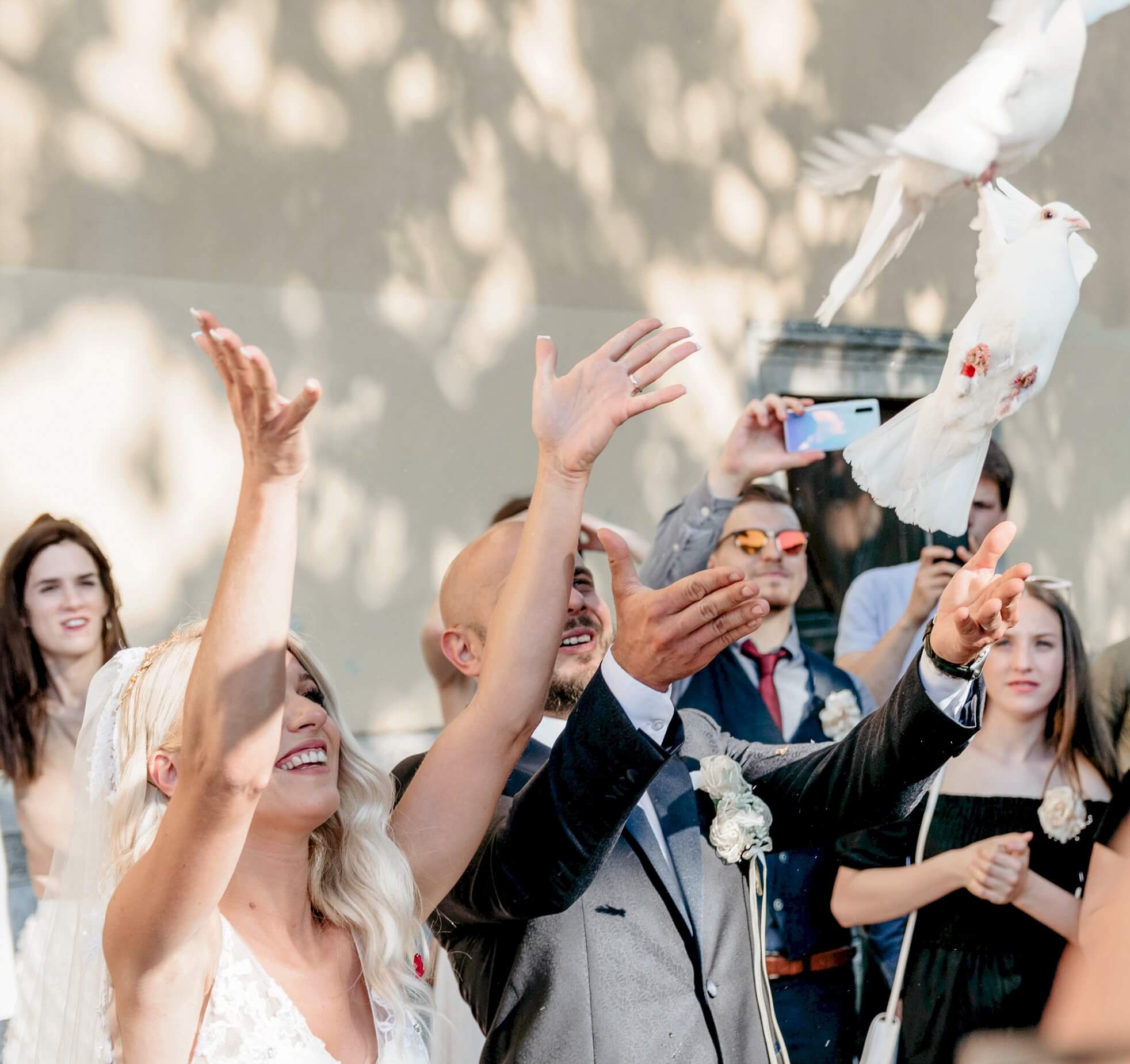 Bride and groom releasing white doves at their wedding, surrounded by smiling guests capturing the moment. Bright, joyful atmosphere.