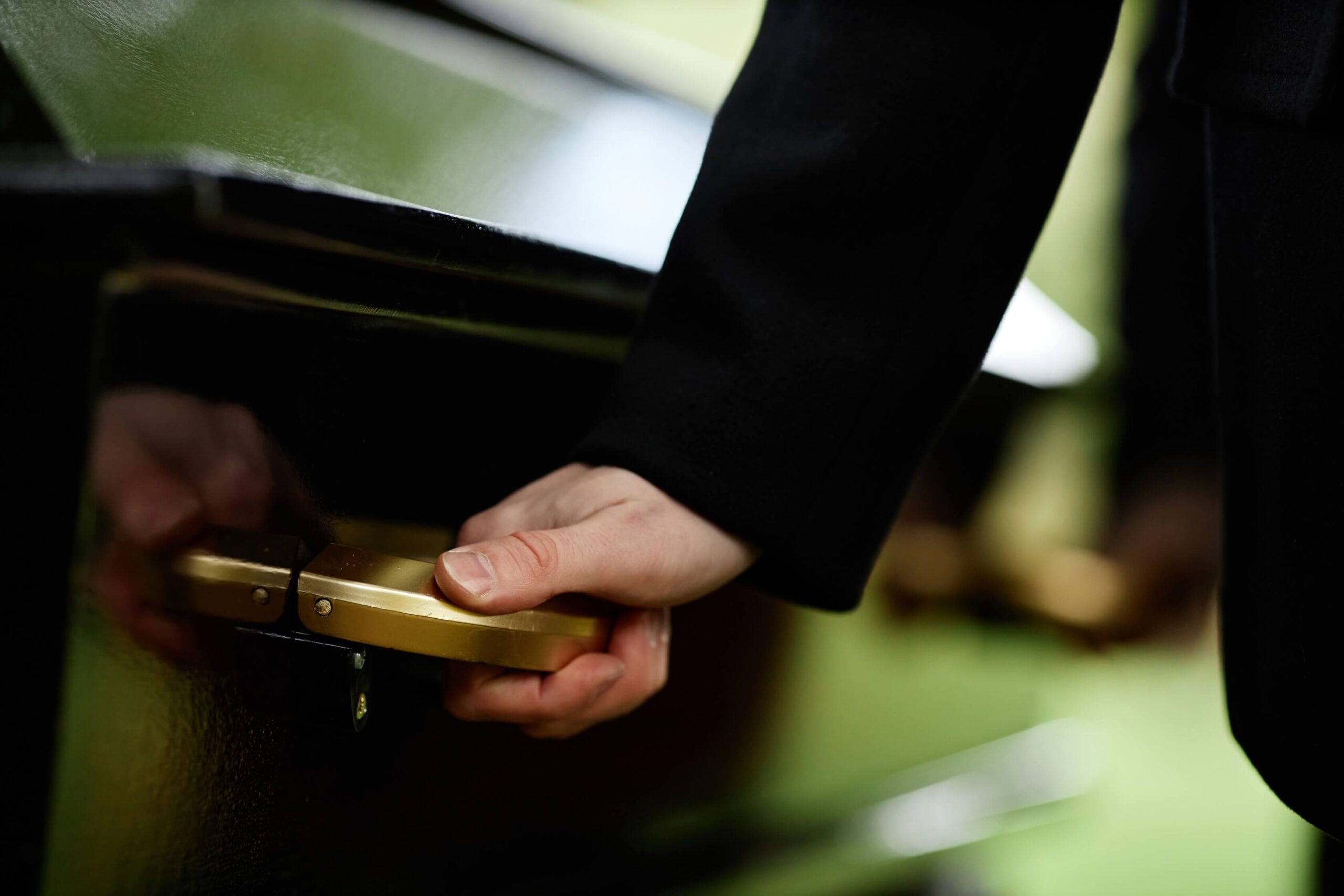 A close-up of a hand gripping a gold-colored handle on a black, glossy surface, possibly a casket. The scene conveys a somber, reflective mood.