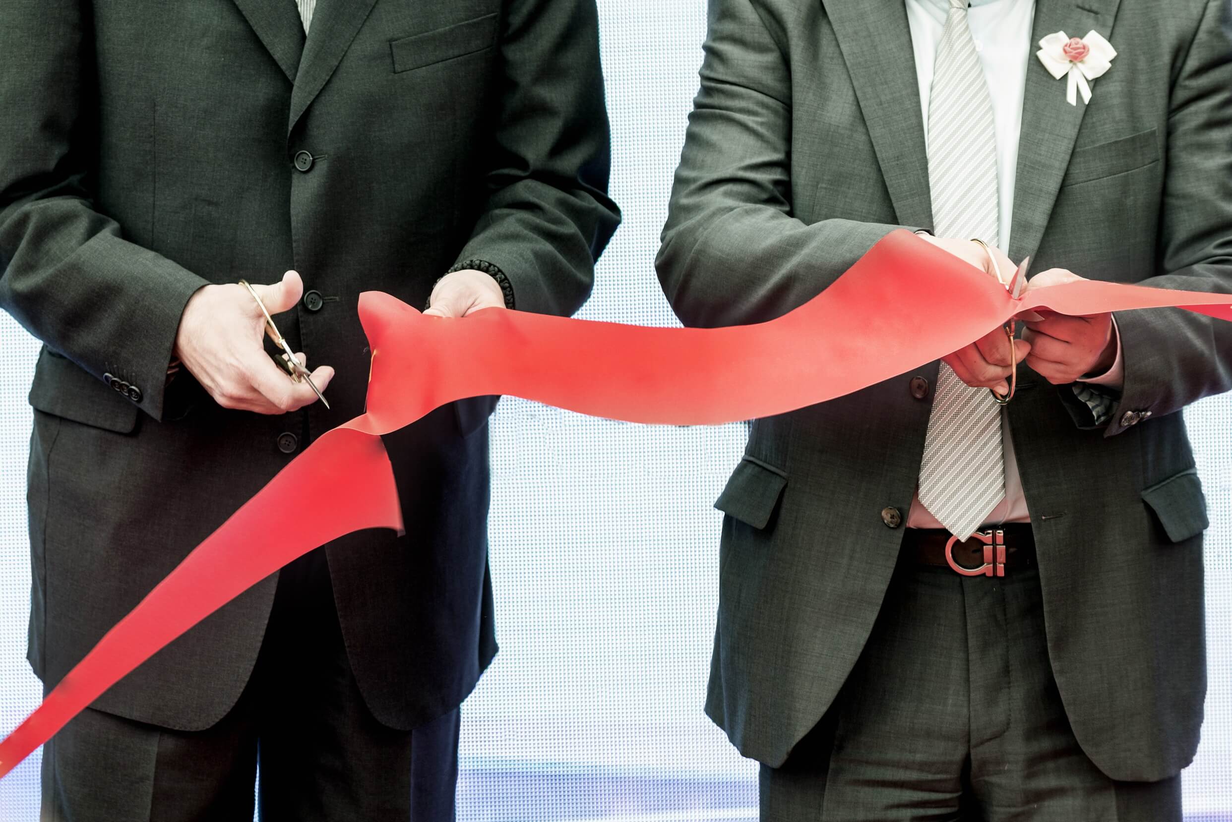 Two individuals in suits cutting a red ribbon with scissors, symbolizing an opening ceremony. The mood is formal and celebratory.